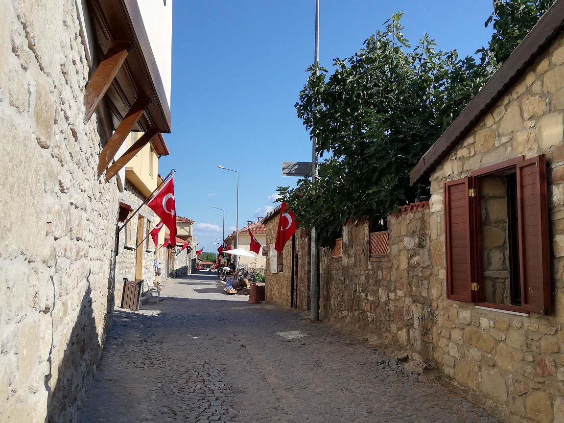 KÖYÜN KADINLARI TEZGAHLARININ BAŞINDA SATIŞ YAPIYOR
Meydanda başlayıp, ara sokağa doğru sıralanan evlerin önünde uzanan dar sokakta, yerlerde tezgah açan köyün kadınları; evlerinde ürettikleri tarhana, erişte, zeytin, zeytin yağı ve bağ-bahçe ürünü domates-biberler, el örgüsü ile hazırladıkları bebek, sabunluk ve yöre için üretilmiş birçok hediyelik eşya satıyorlar. Oturdukları yerden bir yandan da örgü örmeye devam ediyorlar.