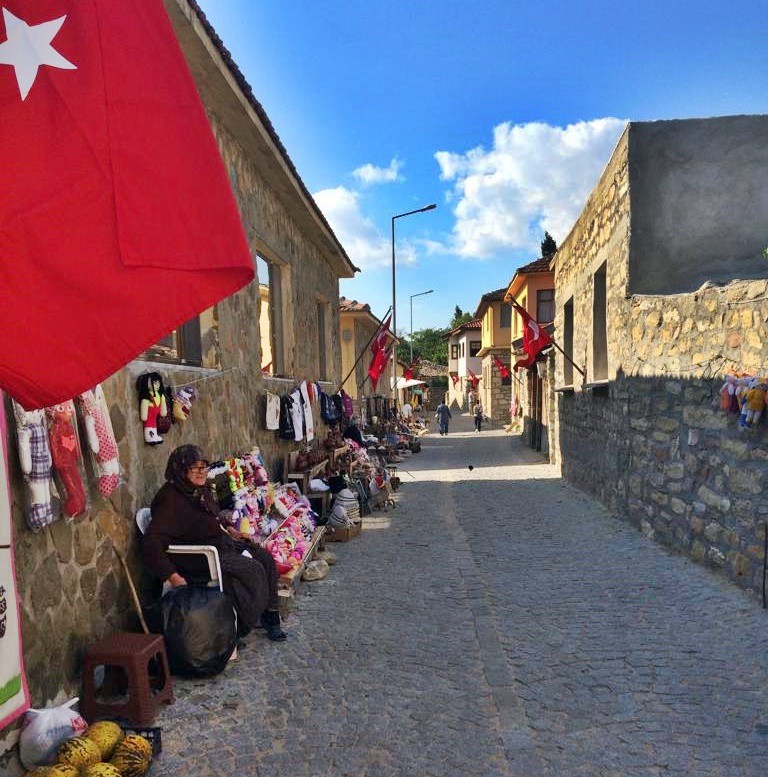 DUVAR BOYU EL İŞİ TEZGAHLARI SIRALIKahvenin karşısında yerel ürünler satan köylü teyzeler sıra sıra dizilmiş. Arkamızda ise dev bir Atatürk posteri asılı. Orada durmuş sanki köye gelenleri karşılıyor.